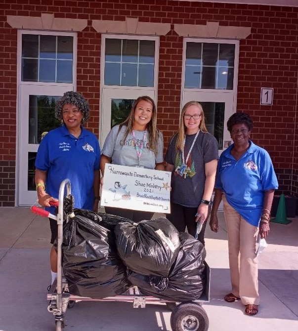 Four women standing with a large check and trash bags outside a brick building.
