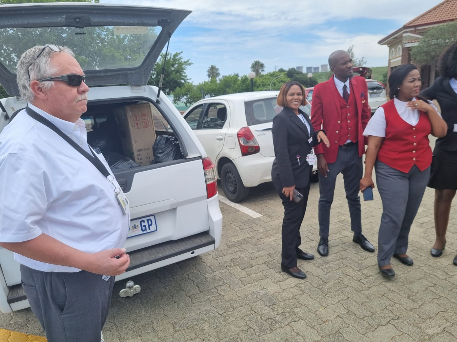 A group of four people standing near parked cars outdoors on a sunny day.