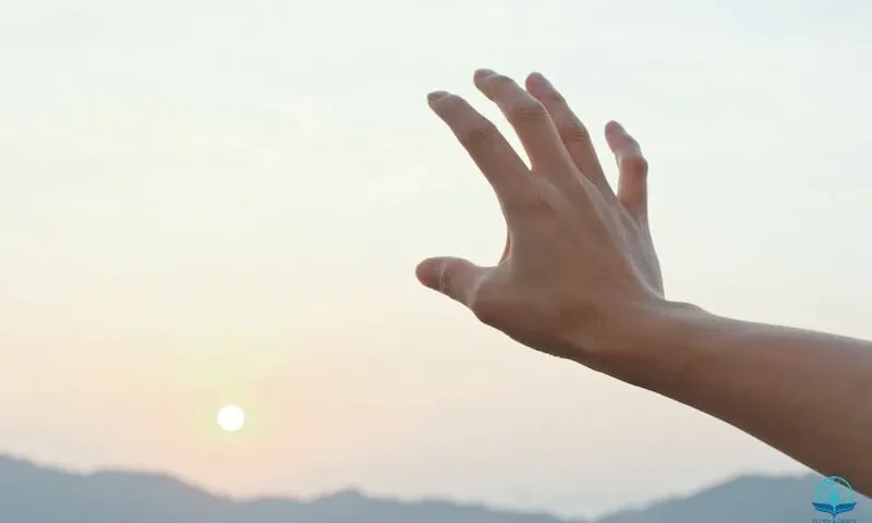 A hand reaching towards the setting sun over distant mountains.